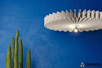 Witte decoratieve hanglamp met bladachtig design tegen blauwe muur, naast groene cactus.