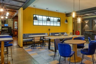 Moderne kantoorlounge met gele muren, blauwe velours stoelen rond houten tafels, grijze zitbanken, hangende verlichting en ho