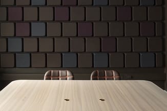 Lichte houten vergadertafel met twee bruine gestoffeerde stoelen tegen een gestructureerde donkerbruine tegelwand.