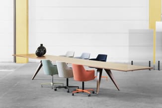 Lange houten vergadertafel met zwart onderstel, omgeven door gekleurde bureaustoelen (groen, grijs, oranje, blauw) in industr