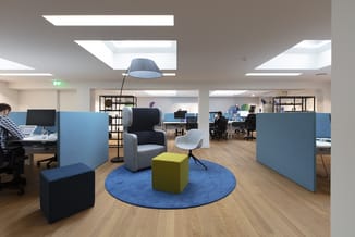 Modern openplan kantoor met blauwe akoestische schermen, grijze fauteuil, gele en donkerblauwe poefs op rond blauw tapijt, ho