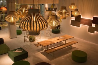Showroom met houten hanglamp, houten picknickbank, groene poefjes en houten wandpanelen in warme verlichting.