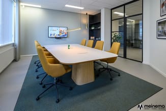Moderne vergaderruimte met witte ovale tafel, gele stoelen met zwarte poten, blauwe tapijt, wandscherm en glaswand.