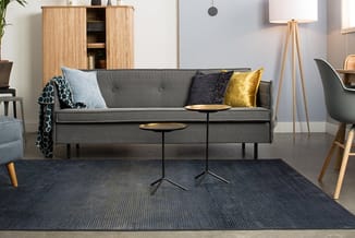 Grijze loungeset in moderne woonkamer met houten kast, gele en blauwe kussens, zwarte tafel en tripod vloerlamp.