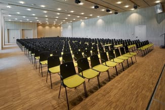 Conferentiezaal met rijen zwarte stoelen met gele zitjes, houten vloer, witte wanden, spotlights in plafond, deuren aan zijka