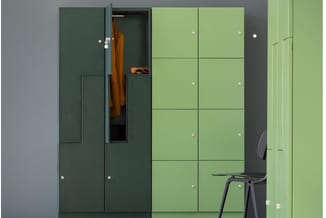 Kastensysteem in groen en donkergroen met open vak waarin oranje kledingstuk hangt, zwarte stoel ervoor, grijze wand.