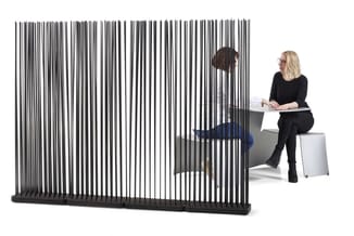 Zwart verticale lamellenafscherming tussen twee personen in kantoorruimte; links figuur achter scherm, rechts zittende vrouw