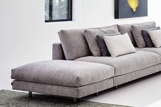 Grijze hoekbank met chaise longue, losse kussens in grijs en ecru, op houten poten in een moderne kantoorruimte.