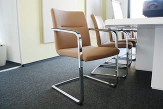 Kantoorruimte met bruine bezoekersstoelen met chromen frames, witte tafel en grijsblauwe vloerbedekking.