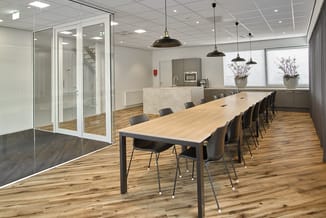 Moderne vergaderruimte met lange houten tafel, zwarte stoelen, glazen wanden, hangende zwarte lampen, keukenzone en houten vl