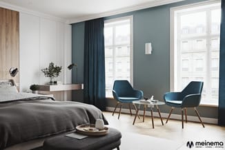 Moderne slaapkamer met grijze bedden, blauwe loungestoelen, houten accenten en teal wandkleur tegen grote ramen.