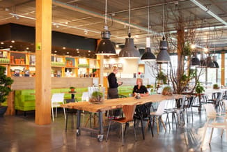 Modern kantoor met houten tafel op wielen, zwarte hanglampen, groene zitbank, gele kastenwand, potplanten en twee personen in