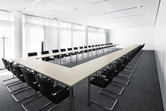 Moderne vergaderruimte met lange witte tafel, zwarte kantoorstoel met chromen frame, grijs tapijt en grote ramen met witte zo