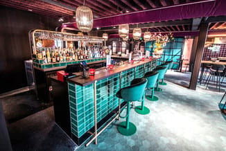 Moderne bar met turquoise tegels, zwarte werkblad, turquoise barkrukken, hangende lampen en achtergrondverlichting in donkere