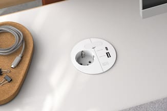 Ronde witte wandcontactdoos in bureauwerk met geïntegreerde EU-stopcontact en twee USB-poorten, geplaatst in wit werkblad.