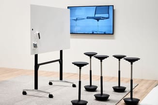 Presentatieruimte met mobiel whiteboard, wandmonteerde TV en vijf zwarte staande zitballen op stalen poten.