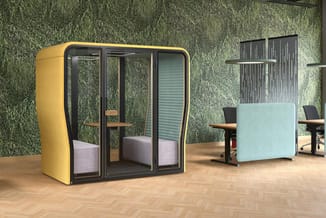 Gele akoestische belcel met glazen deur en roze zitting in kantoorruimte met groene plantenwand en werkplekken.