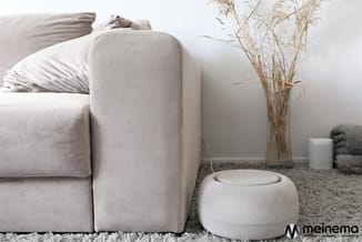 Beige stofbank met hoekdetail naast ronde poef, op grijze vloerkleed met droogbloemen en textielaccessoires.
