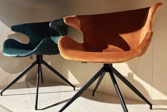 Twee moderne stoelen met draaibare zwarte tripod-poten: één in terracotta velours, één in petrolblauw velours, in showroom te