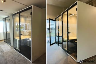 Twee glazen belcellen met zwarte aluminium frames en witte zijpanelen in kantooromgeving, met gele werkbladen en verlichting.