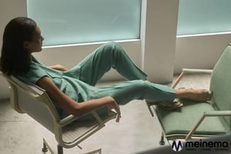 Vrouw zittend in een beige loungestoel met groen kussen in een kantoorruimte met grote ramen en groene akoestische scheidings