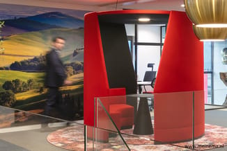 Rode akoestische belcel met zwart interieur in kantoorruimte, met rode fauteuil en bureau, tegen achtergrond met landschapsfo