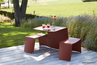 Rood-bruine picknicktafel met bijbehorende banken op houten terras, uitzicht op bloemenweide en grasveld.