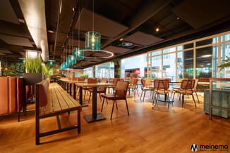 Modern kantoor met houten vloer, zwarte tafels, bruine stoelen, glazen hangende lampen, groene planten en grote ramen.