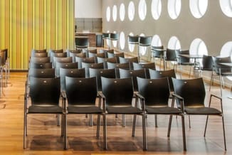Vergaderruimte met zwarte stapelstoelen in rijen, houten vloer, gele gestreepte gordijnen, beige wanden en ronde witte plafon