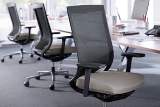 Moderne kantoorwerkplek met zwarte mesh-bureaustoel met beige zitting op voorgrond, achtergrond toont lange vergadertafel met