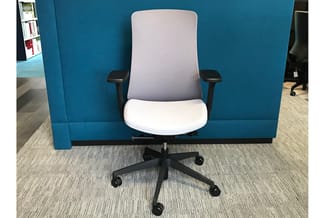 Ergonomische bureaustoel met grijze rugleuning, wit zitgedeelte en zwarte armleuningen tegen blauwe akoestische wand in kanto