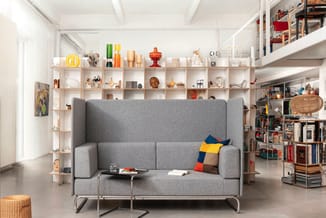 Grijze loungebank met hoge rugkussens op metalen onderstel, in moderne open ruimte met houten wandkasten vol decoratie en boe