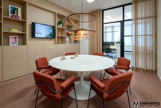 Moderne vergaderruimte met ronde witte tafel, acht rode stoelen, hangende koperen lampen, beige kasten met tv en planken, bei