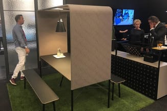 Belcel met beige stofbekleding en zwarte poten in kantoorruimte met kunstgras, werkblad en verlichting. Personen en televisie