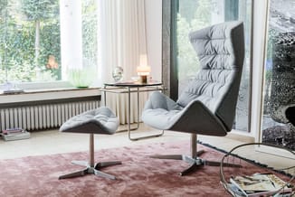 Grijze relaxstoel met voetsteun op wielen in moderne kantooromgeving met raam, zittafel en roze vloerkleed.