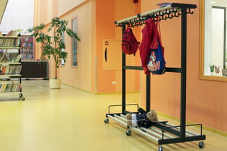 Zwart garderoberek met hangers en wielen in gele schoolgang, rood jasje en blauwe rugzak opgehangen, knuffelbeer eronder.