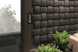 Grijze akoestische wandpanelen in blokpatroon naast een raam met planten op de vensterbank en een SOUNDWAVE-apparaat aan de w