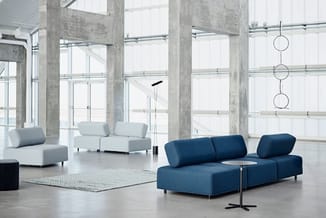 Moderne kantoorruimte met blauwe modulaire bank, lichte stoelen, betonnen zuilen, grote ramen en hangende metalen decoratie.