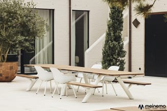 Moderne terrastafels en stoelen in wit en natuurhout in een patio met witte bakstenen gebouw en groene planten.