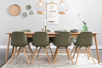 Houten eettafel met vier olijfgroene stoelen, grijze borden, glazen vaas met plant, rieten lampen, pegboard en wanddecoratie