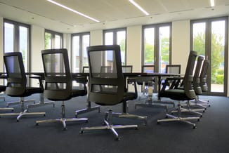 Moderne vergaderruimte met zwarte netkatoelen rond glazen tafel, grote ramen en daglicht, grijs tapijt.