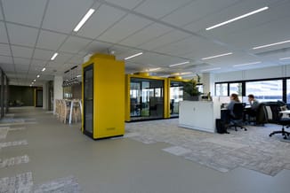 Modern kantoor met gele geluidscabines, werkplekken, witte kasten, grijze vloertegels en plafondverlichting.
