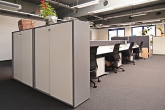Modern kantoor met grijze akoestische schermen, werkplekken met zwarte bureaustoelen, houten accenten en groene planten aan h