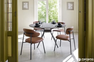 Ronde zwarte eettafel met vier bruine leren stoelen met zwarte metalen poten in lichte ruimte met grote ramen.