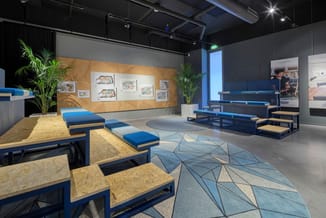 Moderne kantoorruimte met blauwe zitbanken, OSB-tafels, geometrisch patroon vloer, houten wandpanelen, groene planten, uitste