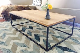Houten salontafel met zwart stalen frame op geometrisch gemusterd tapijt in woonkamer setting, gele bloem in vaas.
