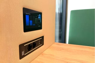 Kantoorwand in beige met ingebouwde zwarte wandcontactdoos en zwart touchscreen-paneel voor vergaderingsbeheer, naast houten