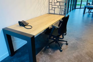 Kantoorwerkplek met zwart onderstel, houten tafelblad, zwarte bureaustoel op wielen, desktop telefoon en open plafond met gro