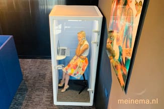 Witte akoestische belcel met glazen deur in kantoor. Woman zit aan bureau met laptop en voert telefoongesprek.
