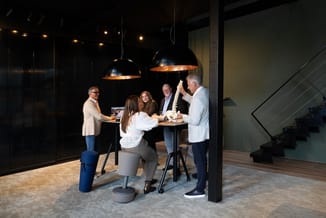 Zes professionals rond een hoge tafel in een modern kantoor met zwarte hanglampem, trap en houten vloer. Één persoon houdt ee